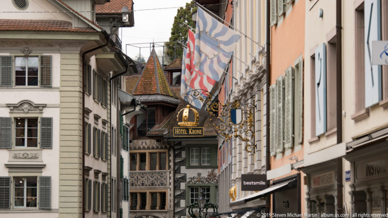 Narrow Lucerne old-town street with shutters, striped flags, ornate Hotel Krone sign, and projecting carved bay.