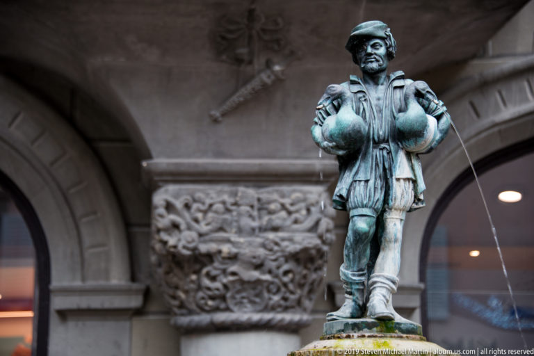 Bronze fountain figure in historic dress standing with hands at hips as water jets arc outward.