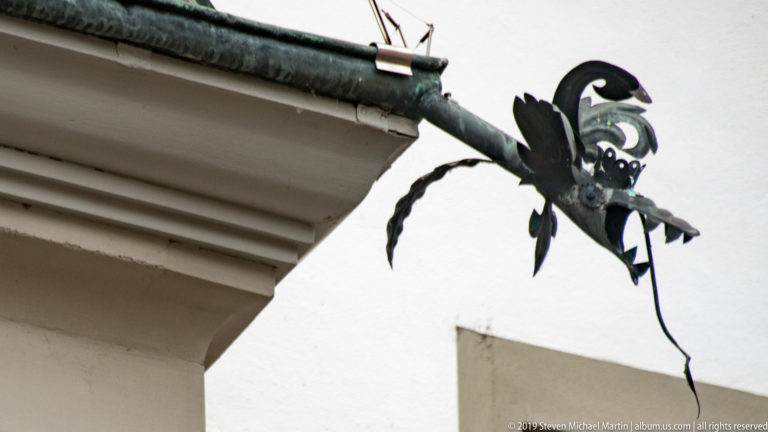 Dragon-shaped dark metal ornament projecting from the end of a roof gutter on a white building.
