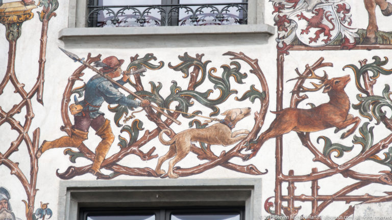 Painted hunting scene on a Lucerne façade with armed hunter, hounds, deer, and scrolling foliage.