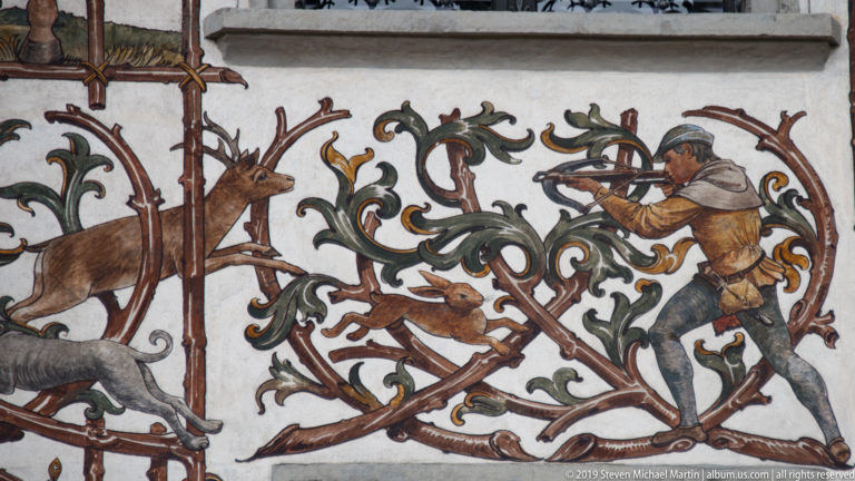 Close section of a painted hunting mural showing a hunter aiming a long firearm at deer among scrolling vines.