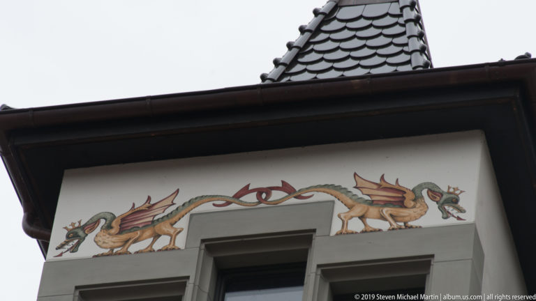Two painted dragons facing an eye-like central motif beneath the roofline of a Lucerne building.