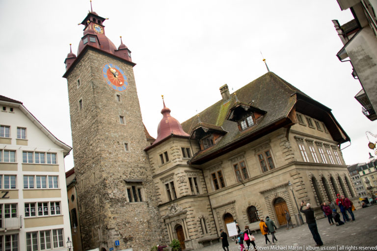 Large painted clock on a square stone tower beside an ornate historic civic building in Lucerne.