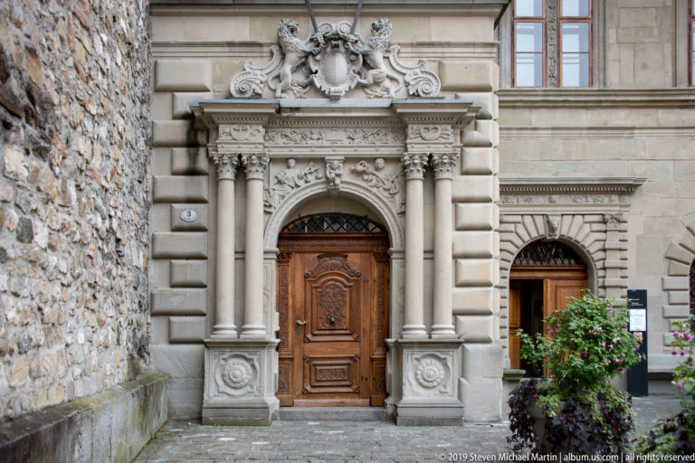 Elaborately carved stone doorway with columns, scrollwork, and a heavy wooden door beside rough masonry.