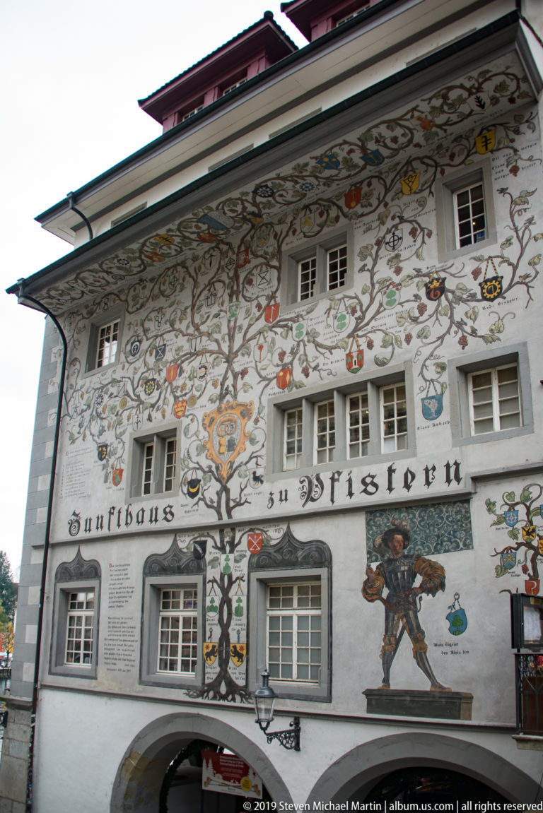 Painted façade of Lucerne’s Zunfthaus zu Pfistern with heraldic shields, scrolling ornament, figures, and identifying inscription.
