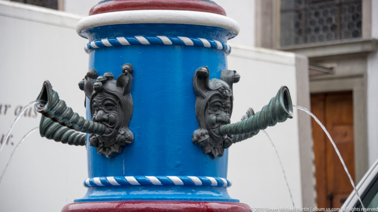 Three grotesque bronze mask spouts projecting from a bright blue fountain column and pouring water.
