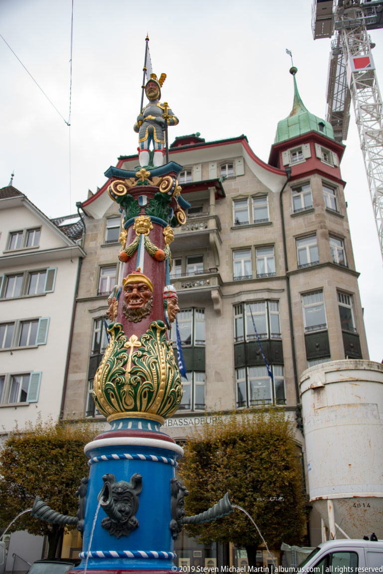 Colorful Lucerne fountain with blue base, green-and-gold sculptural column, masks, heraldic ornament, and crowning figure.