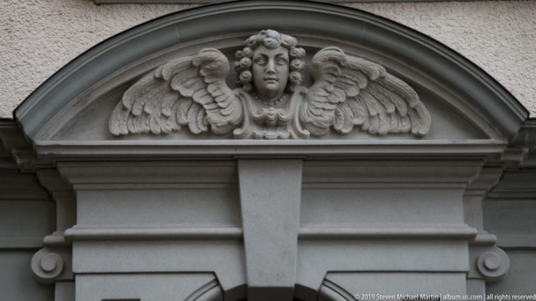 Carved winged angel head filling a curved stone pediment above an architectural opening.