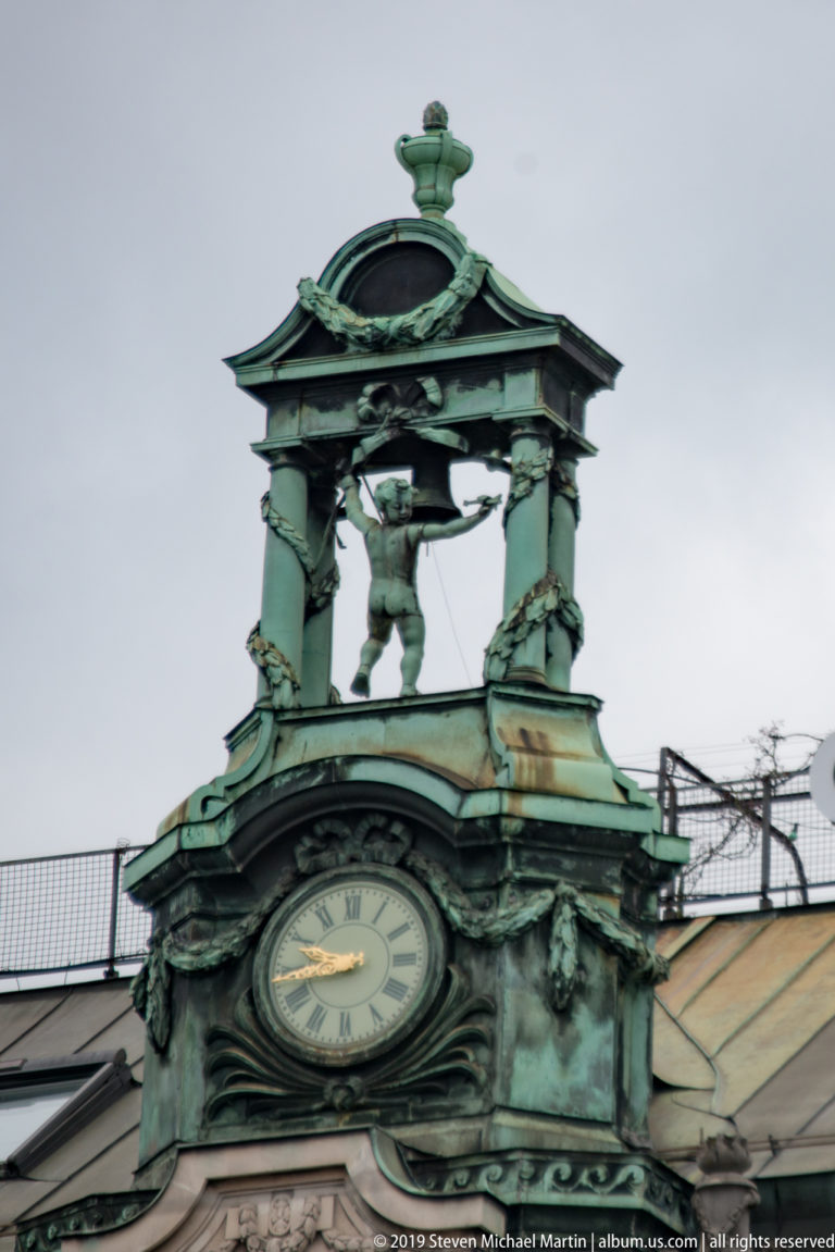 Ornate green-patinated rooftop clock beneath a decorative metal canopy with a small bell and sculptural supports.