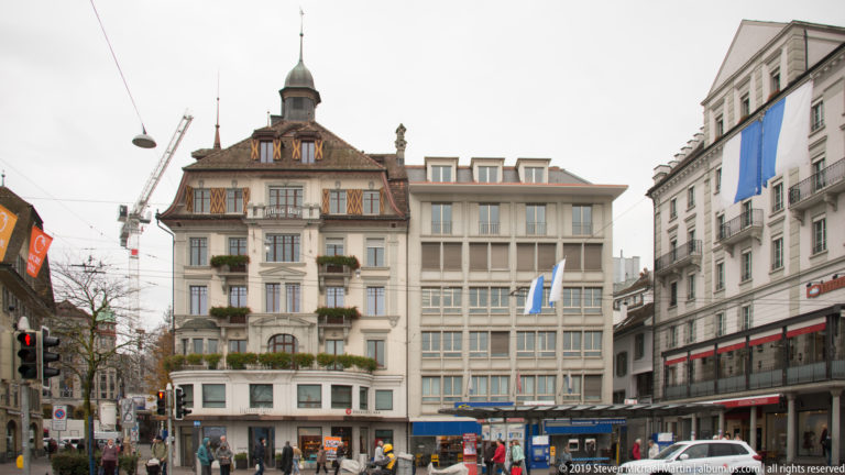 Decorated historic building with steep roof and small turret at the end of a busy central Lucerne street.