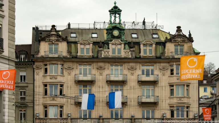 Ornate Hotel Schweizerhof Lucerne façade with balconies, green rooftop clock, and orange Lucerne Festival banners.