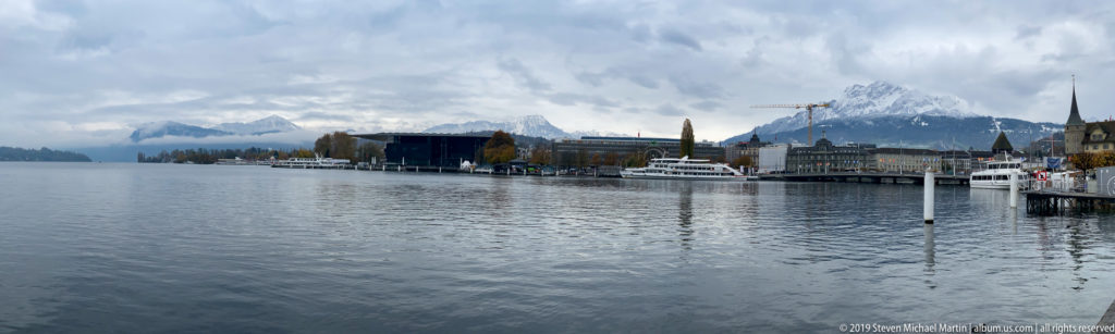 KKL Luzern on the Lake Lucerne shore with excursion boats, quays, and mountains beneath a heavy cloudy sky.