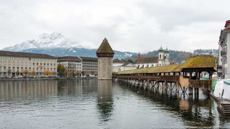 Kapellbrücke leading to the Wasserturm over the Reuss, with Lucerne buildings and snow-covered mountains behind.