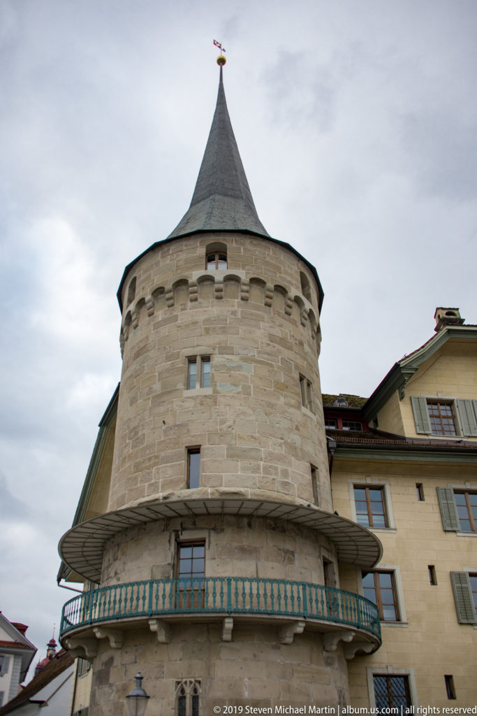 Round stone tower with small openings, projecting gallery, and steep conical roof, viewed from below in Lucerne.