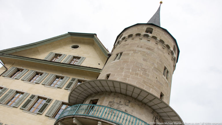 Round stone tower with pointed roof beside a pale shuttered building, seen from a steep upward angle in Lucerne.
