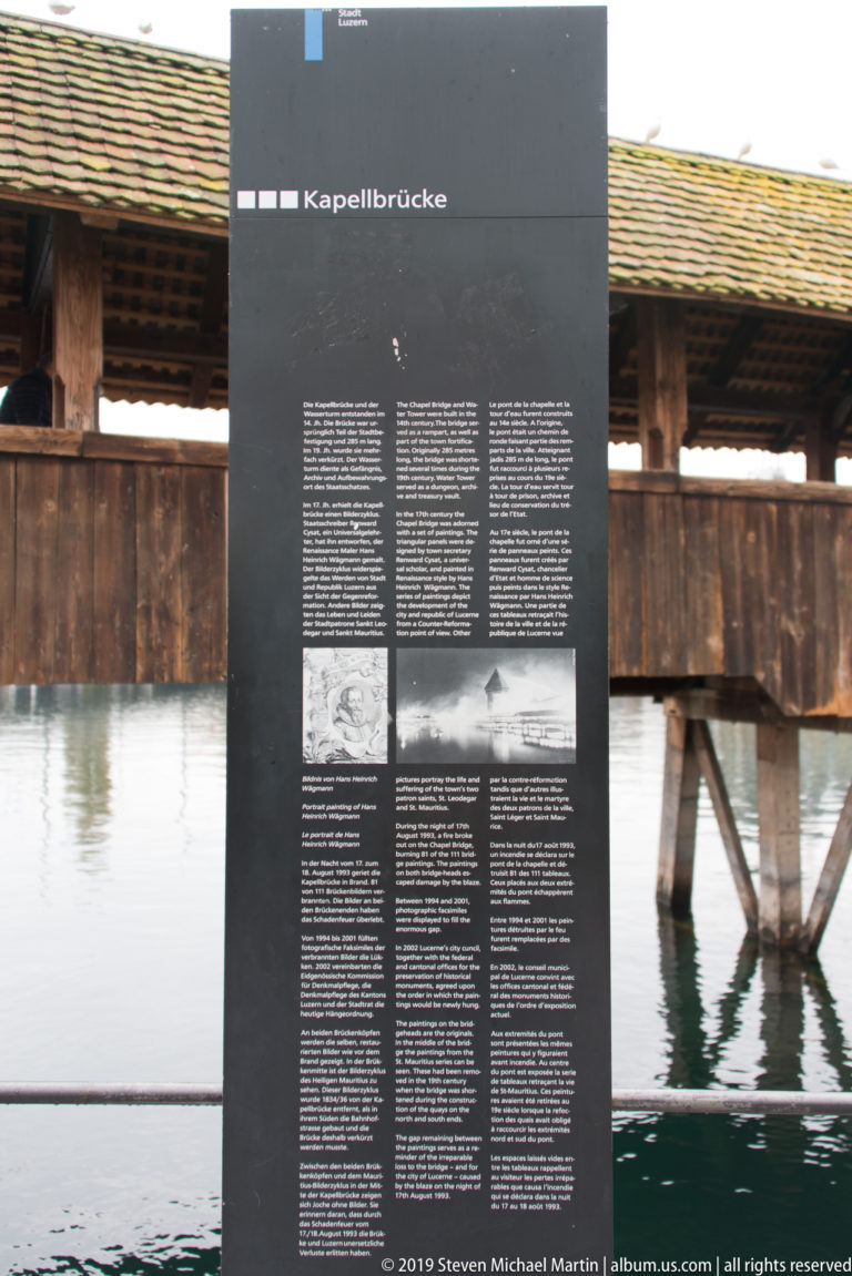 Black interpretive panel labeled Kapellbrücke in front of the covered wooden Chapel Bridge and its supports.