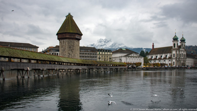 Wasserturm and Kapellbrücke across the Reuss, with Jesuit Church, riverfront buildings, and gulls in the background.