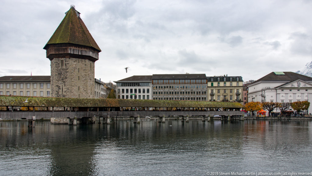 Lucerne Wasserturm beside the Kapellbrücke across the Reuss, with riverfront buildings behind under a cloudy sky.
