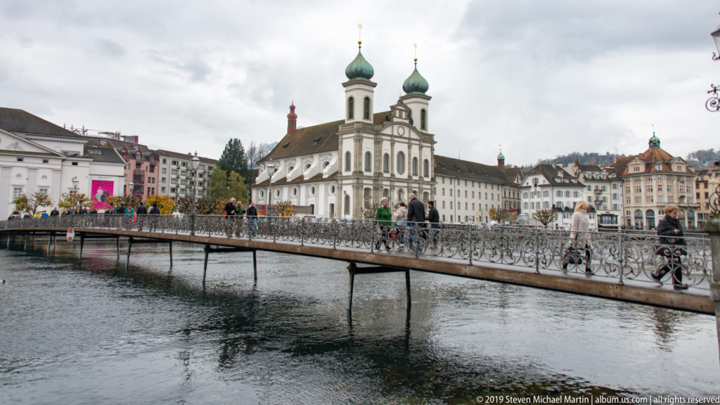 Jesuit Church of St. Francis Xavier across the Reuss, with pedestrians and love locks on a metal footbridge in front.