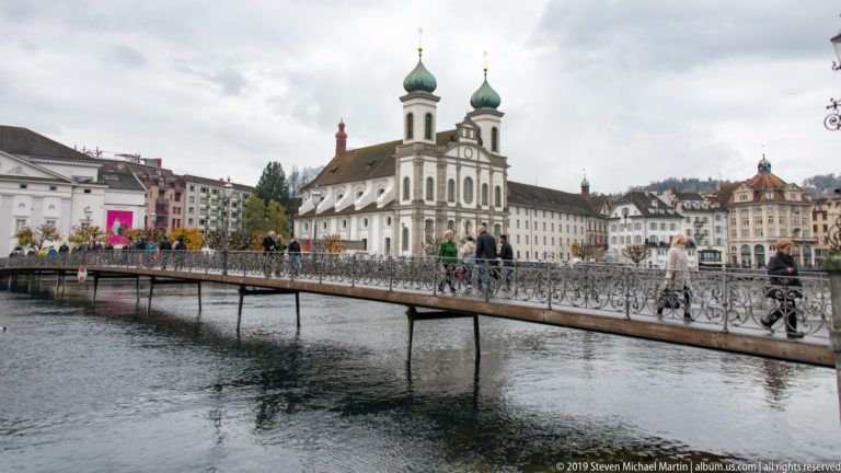 Jesuit Church of St. Francis Xavier across the Reuss, with pedestrians and love locks on a metal footbridge in front.