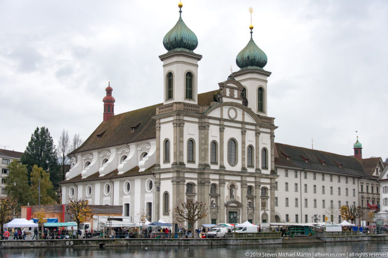 Baroque Jesuit Church of St. Francis Xavier beside the Reuss, with twin onion-domed towers and pale stucco façade.