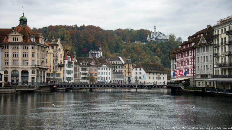 Reuss River through Lucerne with historic waterfront buildings and Château Gütsch visible on the wooded hill beyond.
