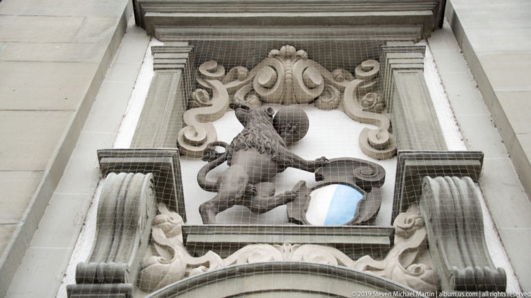 Carved stone lion and shield framed by scrollwork above an arch, with protective netting across the architectural detail.