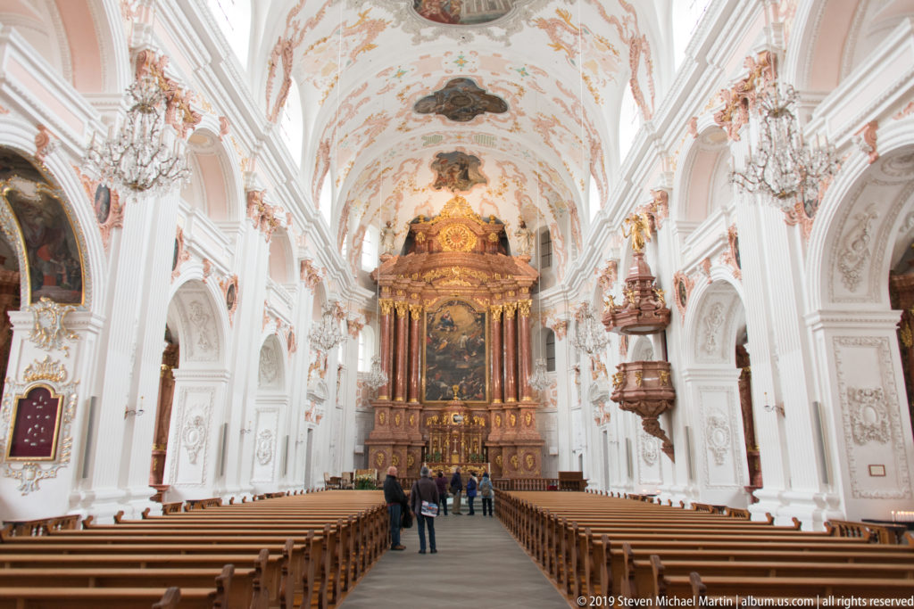 Central nave of Lucerne Jesuit Church with wooden pews, white stucco walls, frescoed vault, chandeliers, and high altar.