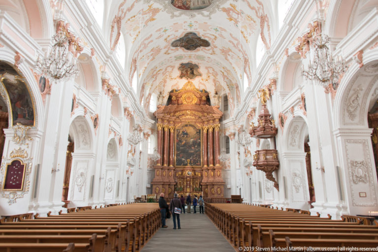 Central nave of Lucerne Jesuit Church with wooden pews, white stucco walls, frescoed vault, chandeliers, and high altar.