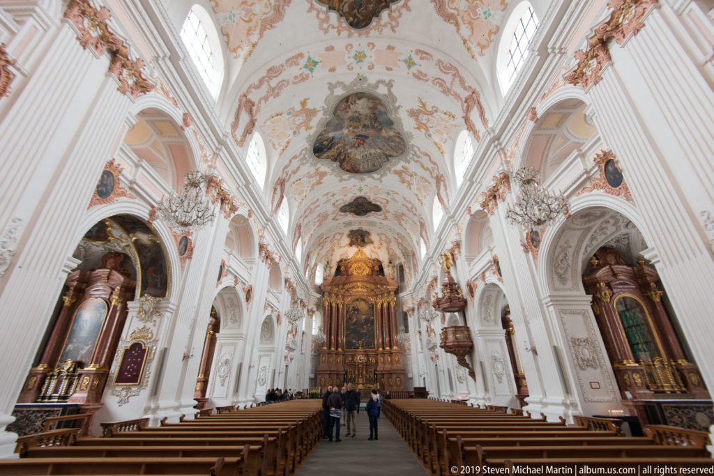 Jesuit Church nave with colorful side altars, white stucco arches, frescoed ceiling, and high altar centered beyond.