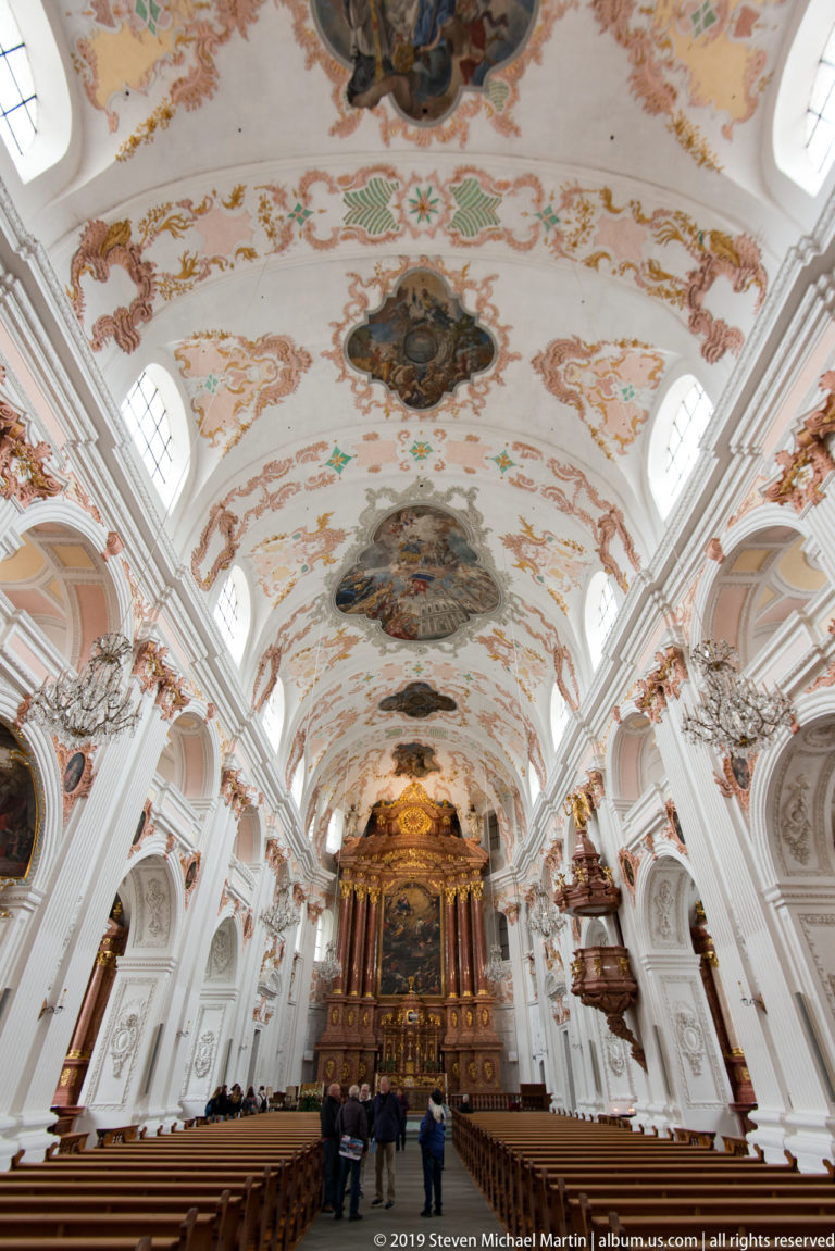 Upward view along the Jesuit Church nave showing fresco panels, ornate stucco, clerestory windows, and chandeliers.