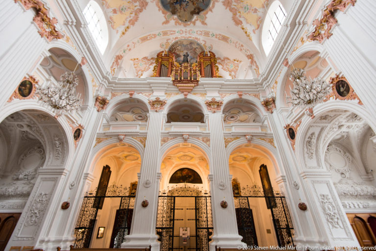 Rear gallery of Lucerne Jesuit Church with pipe organ above arched openings, white pilasters, chandeliers, and iron gates.