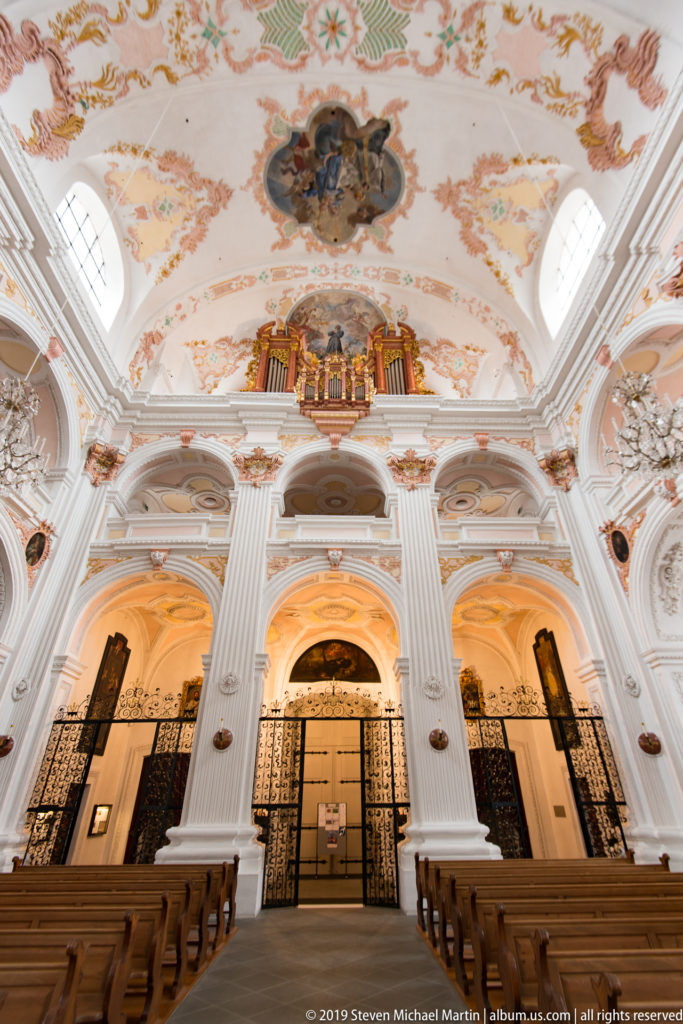 Jesuit Church rear gallery seen over pews, with central pipe organ, white pilasters, arched bays, and ceiling frescoes.