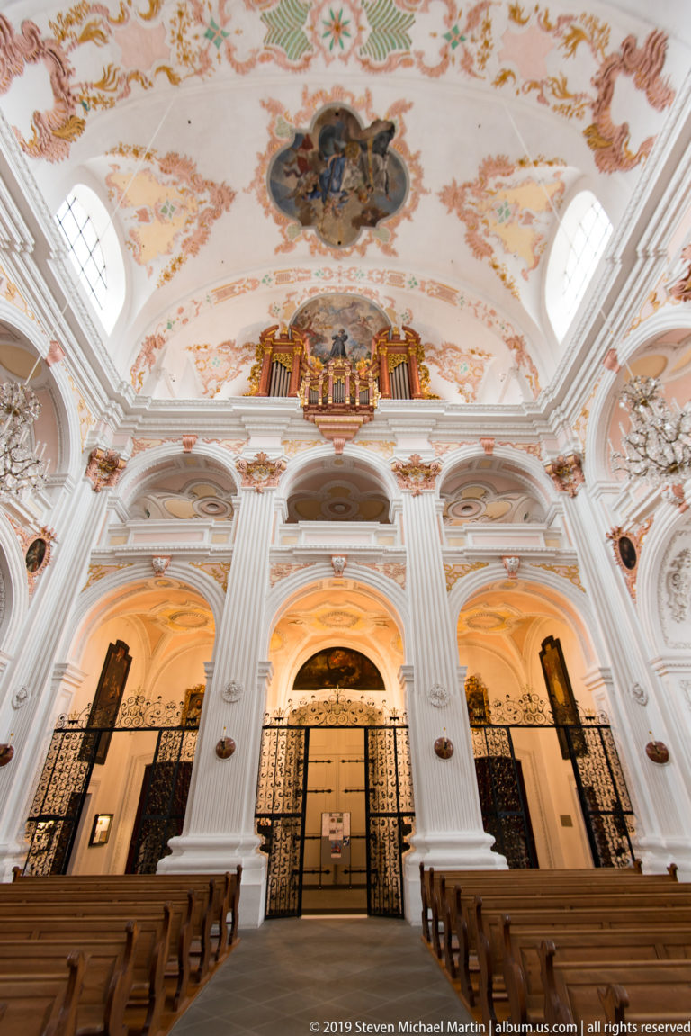 Jesuit Church rear gallery seen over pews, with central pipe organ, white pilasters, arched bays, and ceiling frescoes.
