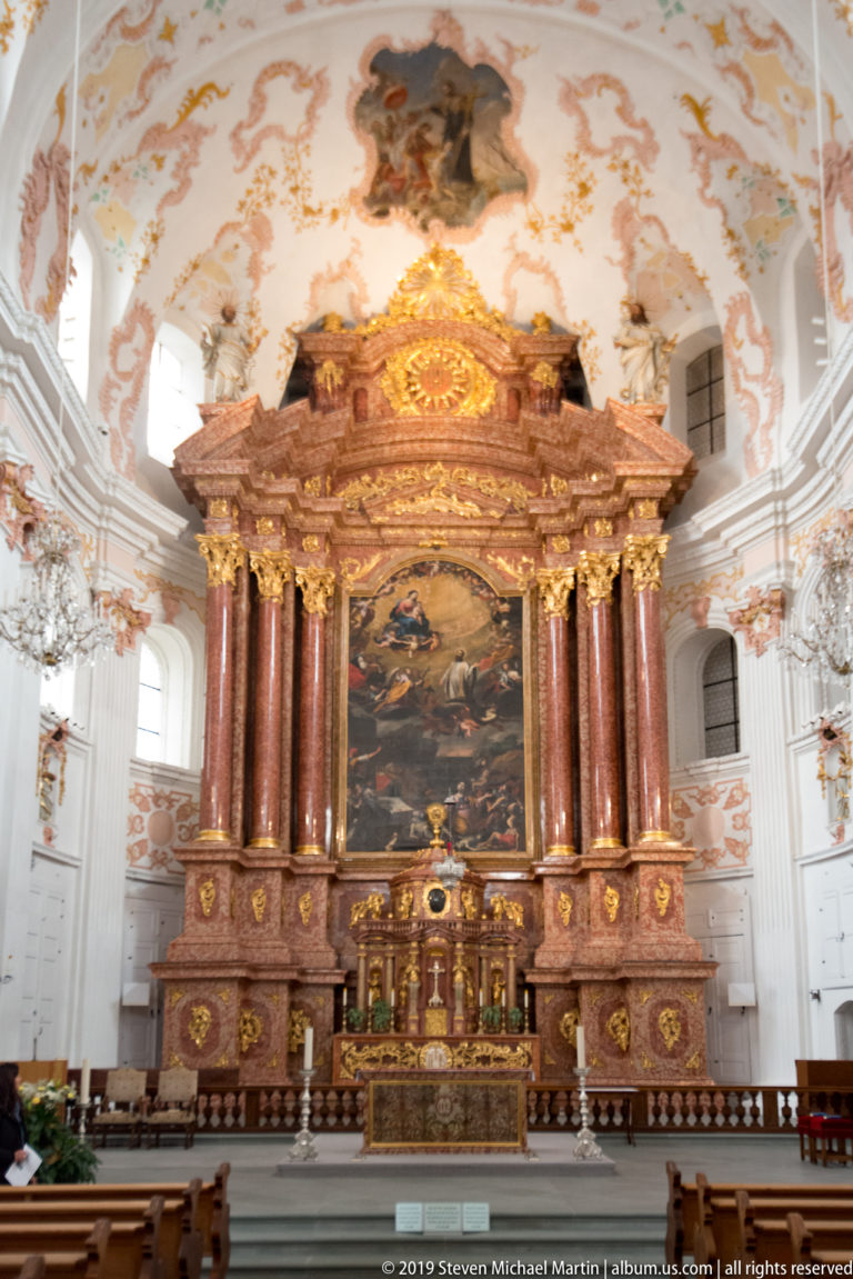 Large Baroque high altar with reddish columns, gilded ornament, central painting, and pale frescoed church interior around it.