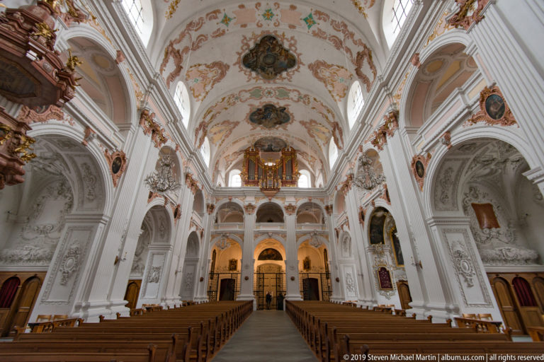 Jesuit Church nave viewed toward the rear organ gallery, with pews, white arches, frescoes, chandeliers, and side altars.