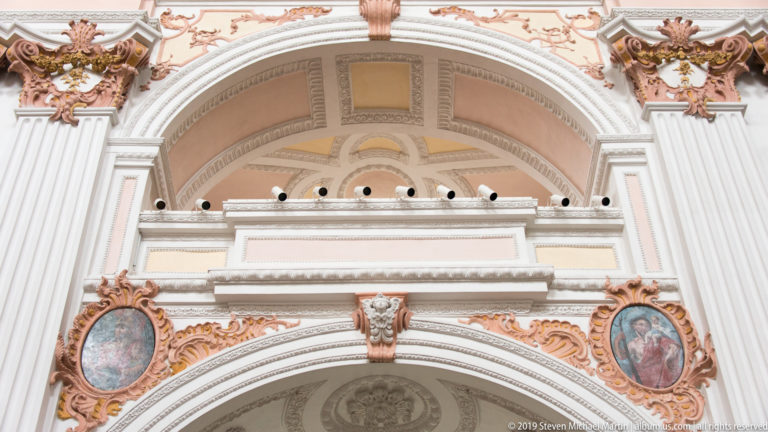 White and peach Baroque arch with ornate stucco, two oval devotional paintings, layered moldings, and small sculptural details.