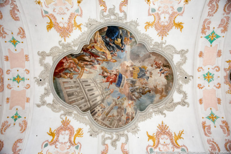 Shaped Baroque ceiling fresco with many figures, clouds, architectural illusion, and an ornate white stucco frame.