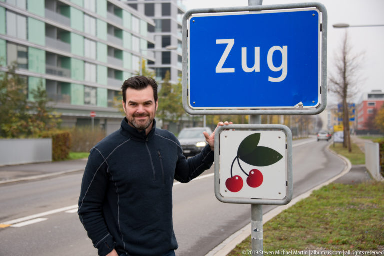 Man standing beside a blue Zug road sign with a red-cherry emblem on a city street.