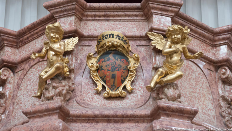 Two gilded winged putti beside a crowned painted shield on reddish stone with carved Baroque scrollwork.