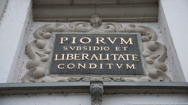 Stone-framed black plaque with gilded Latin inscription PIORVM SVBSIDIO ET LIBERALITATE CONDITVM above an architectural opening.