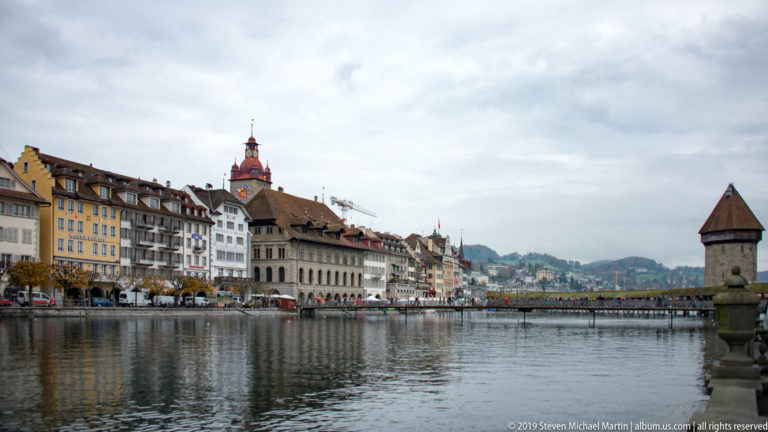 Lucerne’s Reuss waterfront with historic façades, Rathaus roofline, a bridge across the river, and a stone tower at right.
