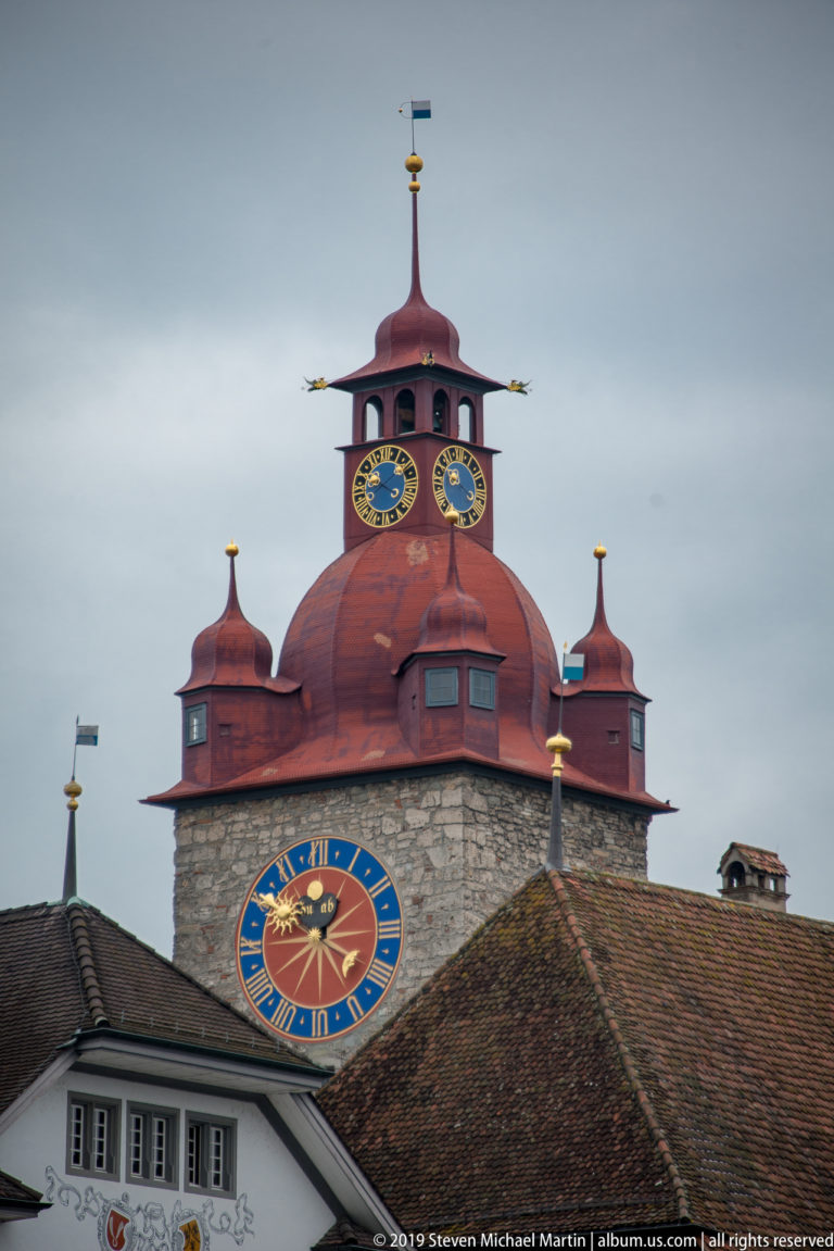 Zytturm on Lucerne’s Musegg Wall with red roof, stone masonry, and two colorful clock faces above tiled rooftops.