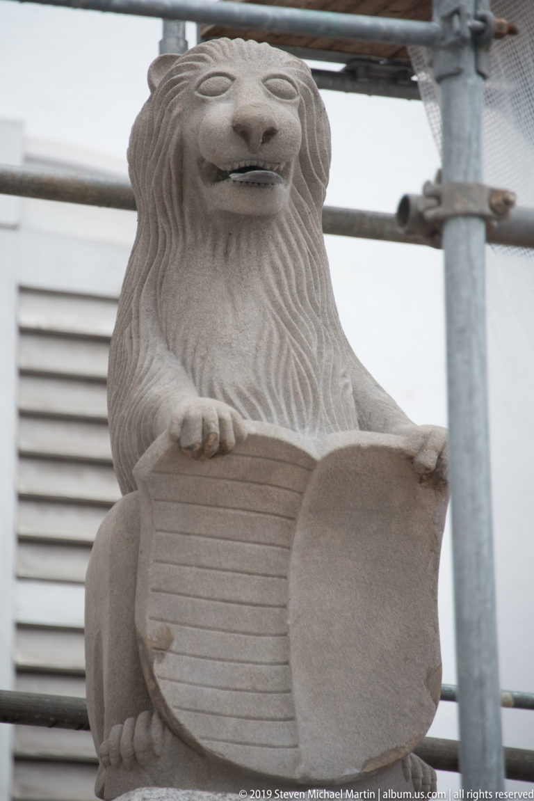 Seated carved stone lion holding a shield, with scaffolding and conservation structures visible around the sculpture.