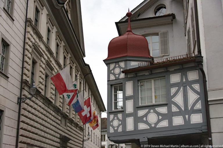 Gray-and-white decorated projecting corner bay with red onion-shaped roof and flags on a historic Lucerne street.