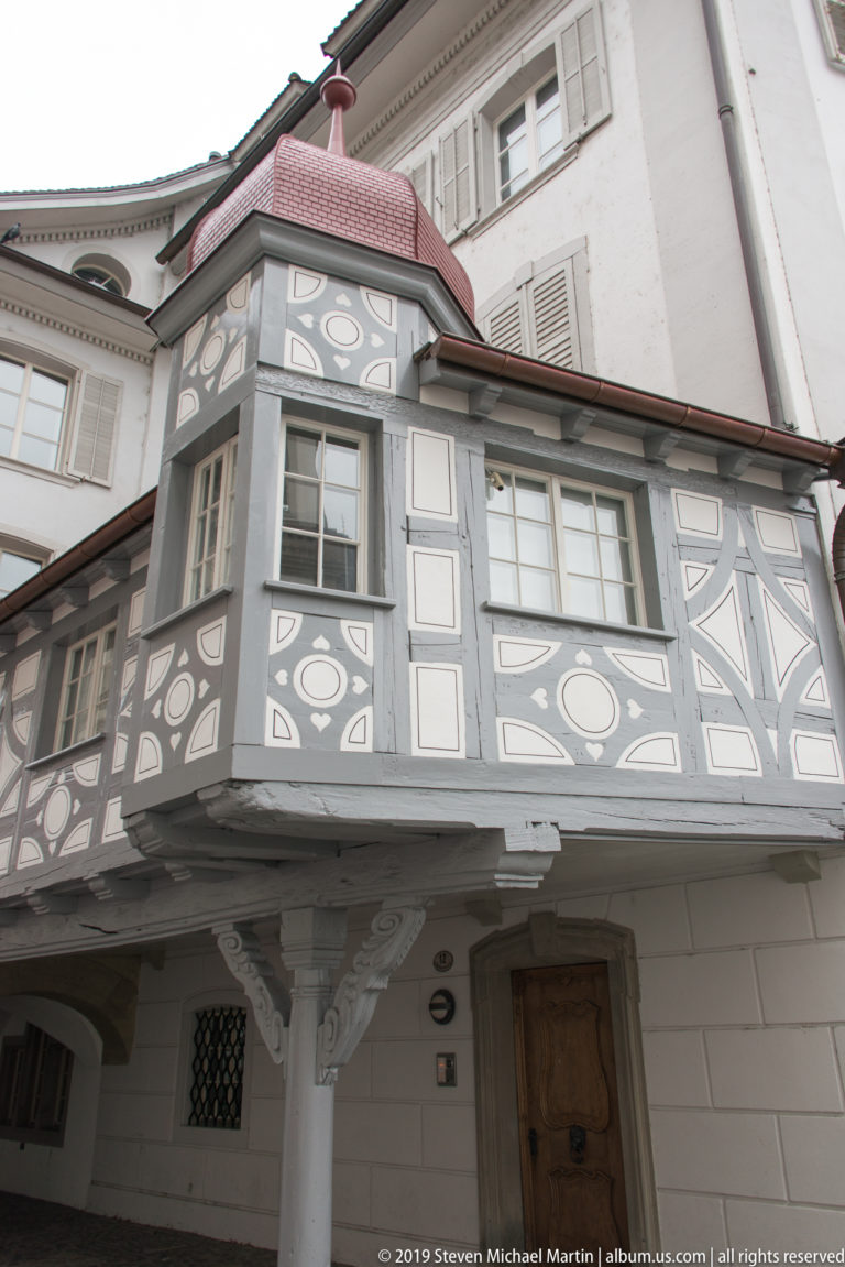 Low-angle view of a painted gray-and-white oriel supported by a column, with windows and a curved red roof.