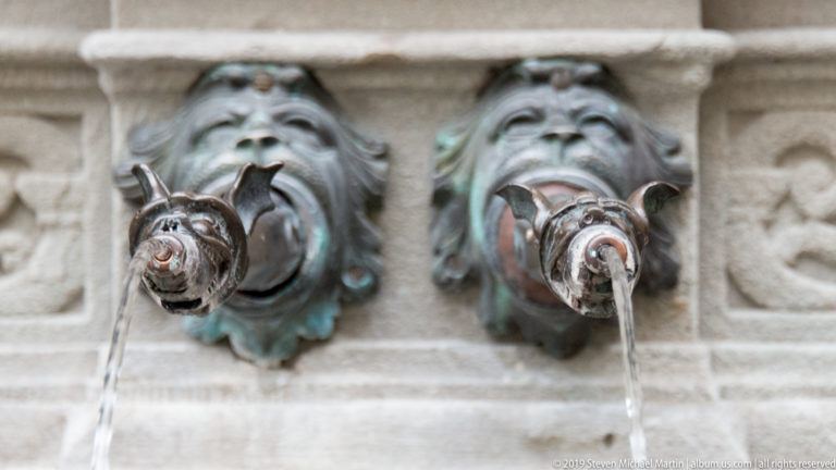 Two patinated bronze lion-head spouts pouring water from a pale stone fountain wall.
