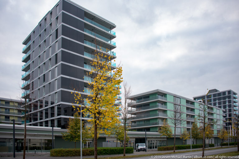 Modern gray and glass apartment or office blocks along a landscaped Lucerne street with yellow-leaved autumn trees.