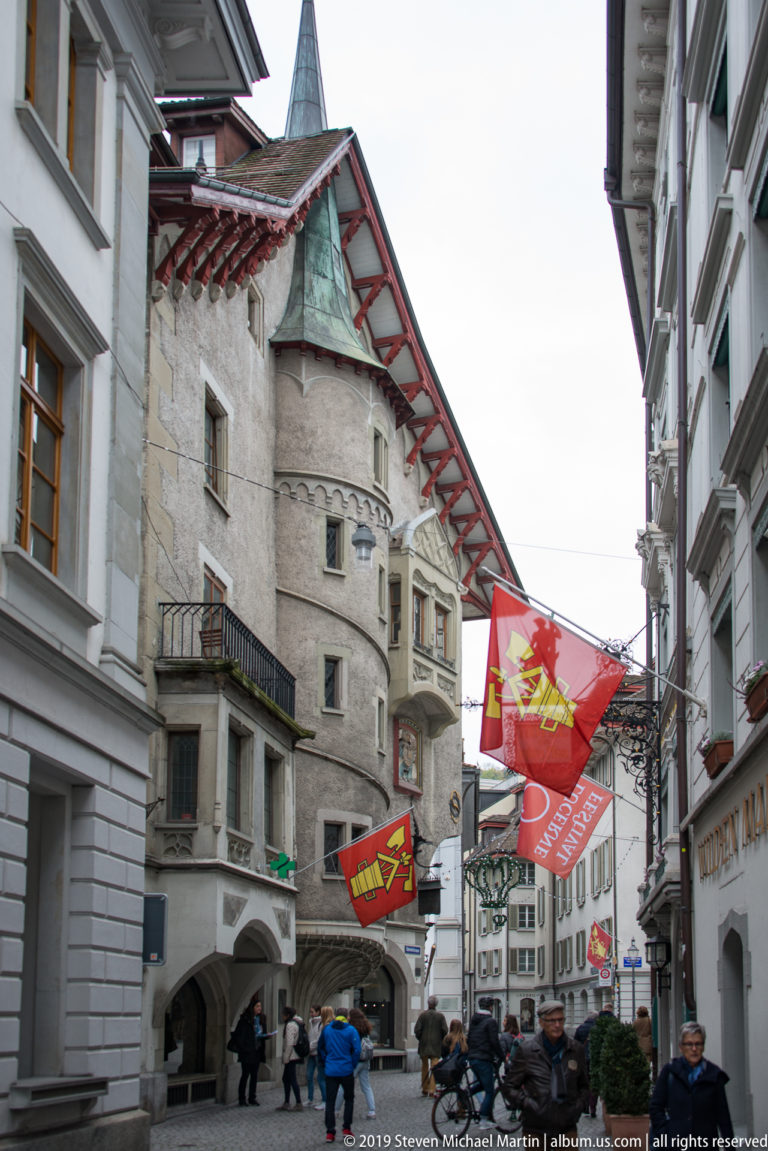 Historic Lucerne street with flags, pedestrians, and a large old corner building with projecting eaves and arched openings.