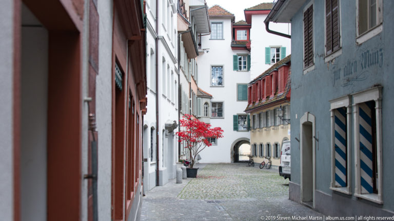 Narrow cobbled Lucerne lane lined with historic houses, shutters, flowers, and a small arched passage.